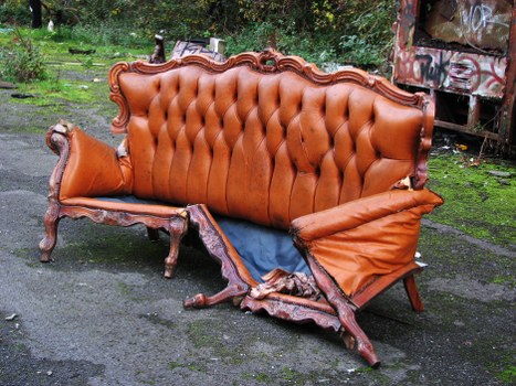 A vintage-style sofa with orange-brown, tufted leather upholstery featuring a carved wooden frame and ornate detailing rests on a paved driveway, partially damaged with a torn section on the seat cushion and a loose armrest. The sofa is positioned outdoors on a rough asphalt surface, with a background of greenery and trees, and a wooden or metal structure visible in the top right corner. The damaged fabric and exposed cushioning indicate it is being prepared for rubbish removal, likely facilitated by Commercial Waste Sudbury, a company offering rubbish clearance services in the Sudbury area. Natural light highlights the texture of the worn leather and intricate woodwork, emphasizing the aged condition of the furniture in an outdoor setting consistent with waste collection activities.
