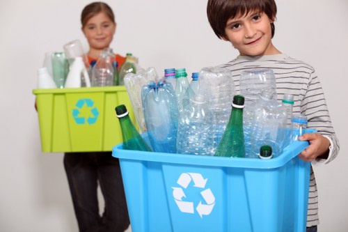 A young boy and girl holding large plastic recycling bins filled with empty bottles and containers. The boy in the foreground is smiling and wearing a grey and white striped shirt, with a blue recycling bin containing various clear plastic bottles, some with green caps and others without. Behind him, the girl is slightly out of focus, holding a green recycling bin with a similar collection of bottles, including clear and green plastic types. The background is plain and white, emphasizing the focus on the bins and their contents. The scene highlights the act of recycling and waste separation, suitable for promoting rubbish removal and recycling services in Sudbury, with a professional and neutral tone consistent with waste management topics.