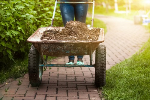 A person wearing blue jeans and footwear seen from the waist down is pushing a wheelbarrow filled with dark, moist soil along a brick-paved pathway. The wheelbarrow has a metal frame with two black rubber wheels, and its tray is weathered with a reddish-brown finish, containing a large pile of soil that appears freshly dug. Surrounding the pathway are lush green bushes and grass, with a well-maintained garden on the right side, indicating an outdoor residential or garden setting. Bright natural lighting suggests daytime, emphasizing the earthy tones of the soil and the greenery. The scene aligns with garden waste clearance and rubbish removal services provided by Commercial Waste Sudbury, serving local homeowners in Sudbury and surrounding areas in Suffolk.