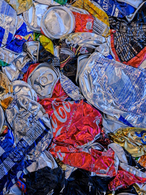 A large collection of mixed household waste and recycling, including black and black-and-white plastic bags, cardboard boxes, paper, and miscellaneous packaging, piled next to a metal recycling bin la