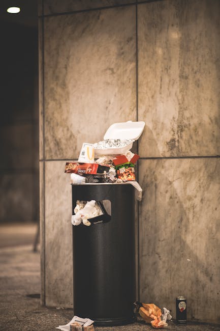 A black, cylindrical litter bin made of metal, positioned against a large, beige stone or concrete wall with visible horizontal and vertical seams. The bin's lid is open, revealing used paper wrap, fo