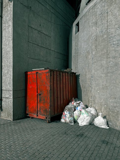 A black, cylindrical litter bin made of metal, positioned against a large, beige stone or concrete wall with visible horizontal and vertical seams. The bin's lid is open, revealing used paper wrap, fo