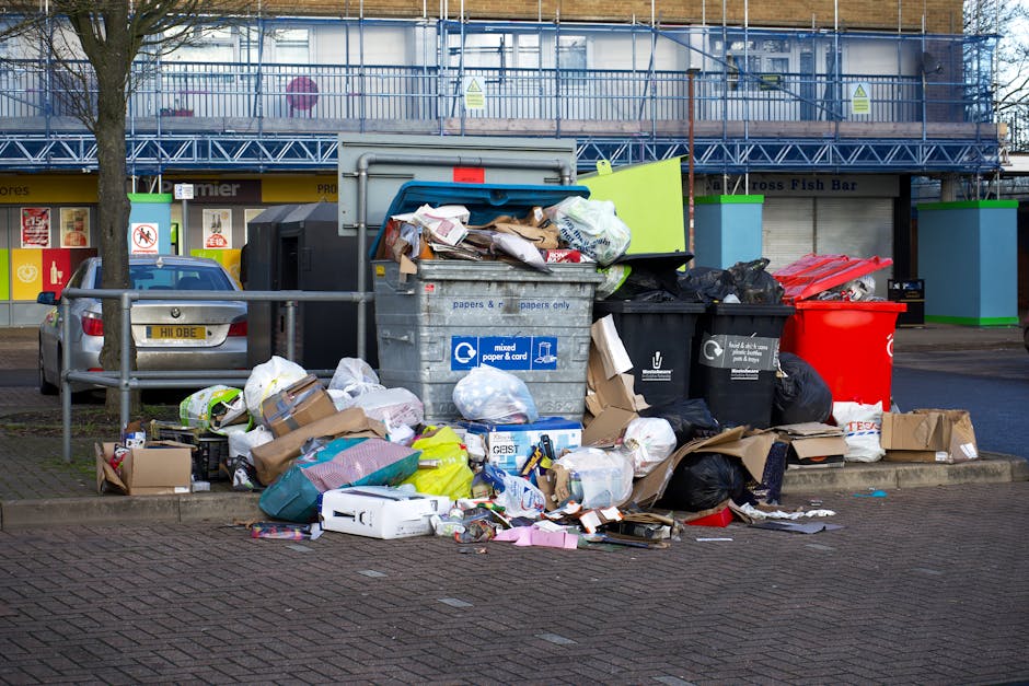 A photograph of a cluttered driveway or outdoor area showing a large pile of mixed waste materials including plastic bags, cardboard boxes, wooden pallets, and household rubbish. The waste appears to
