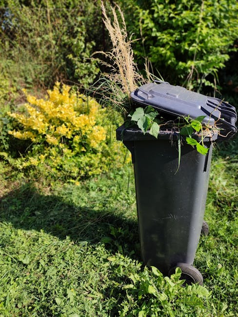 A close-up view of a light grey plastic wheelbarrow with a black rubber wheel, filled with freshly cut green garden waste, including grass, weeds, and small plants, set on a lush green lawn. In the ba